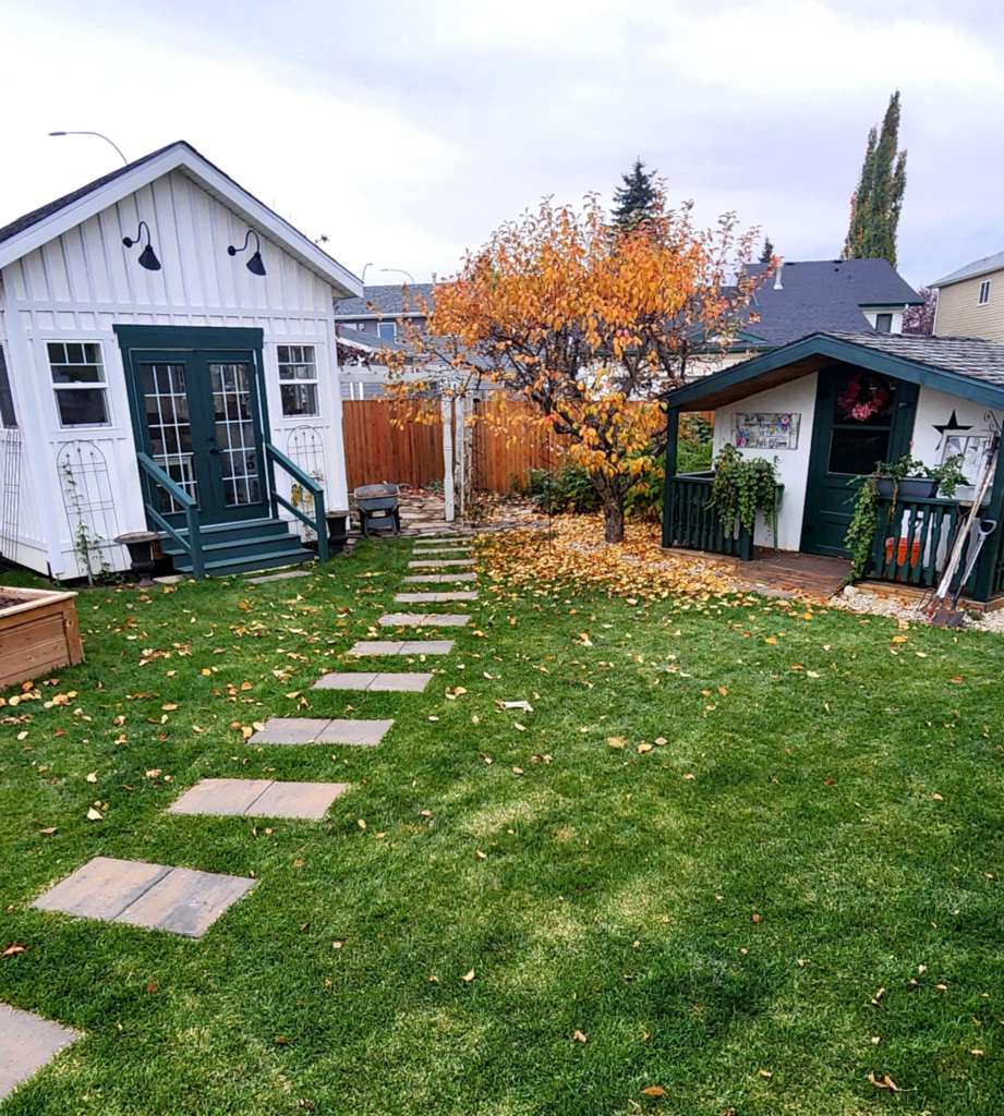 A well-maintained backyard featuring two small sheds, a stone pathway leading to them, and an autumn tree with yellow leaves.