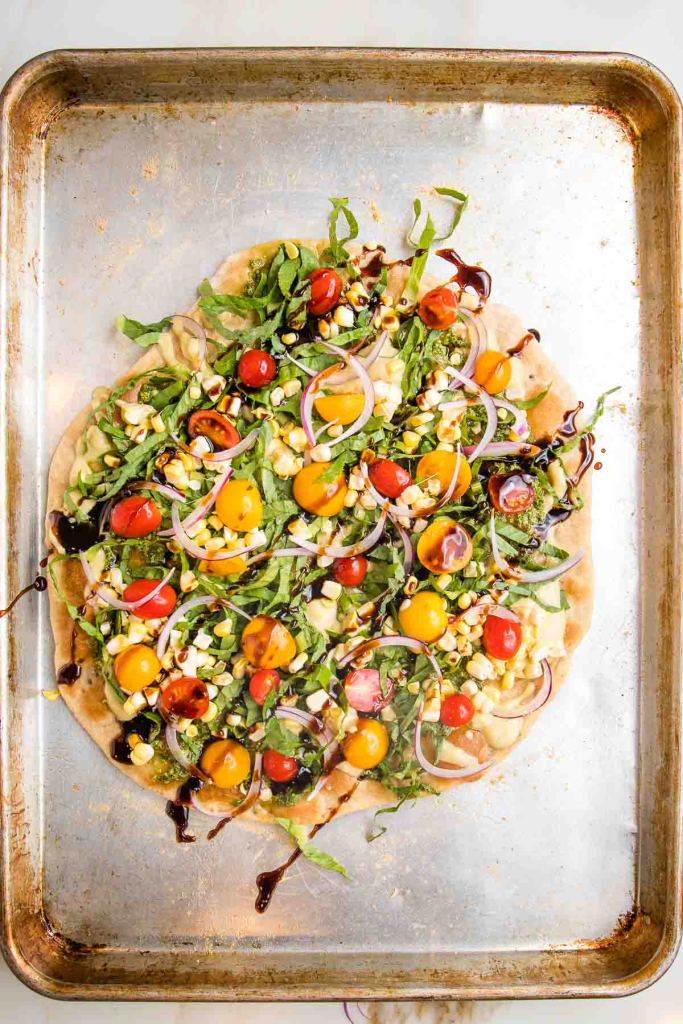 A flatbread pizza topped with tomatoes, onions, corn, arugula, and a drizzle of balsamic reduction. 