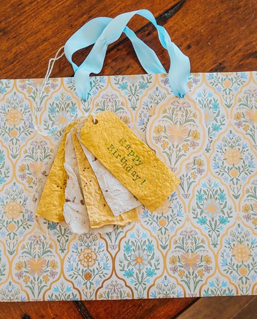 A collection of handmade plantable gift tags in yellow and white, featuring the greeting 'Happy Birthday!' The tags are resting on a decorative floral paper background with a light blue ribbon intertwined.
