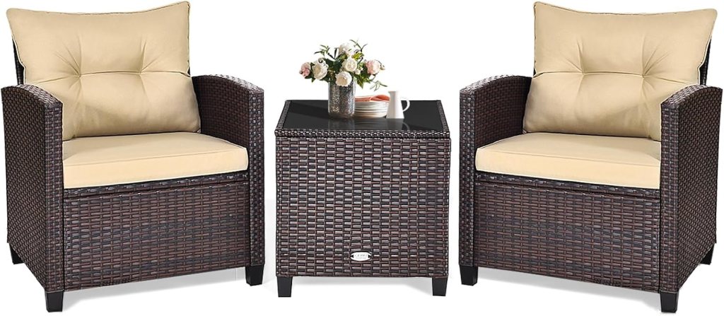 A set of two wicker armchairs with cream cushions and a matching wicker side table, featuring a small vase of flowers and a mug on top.