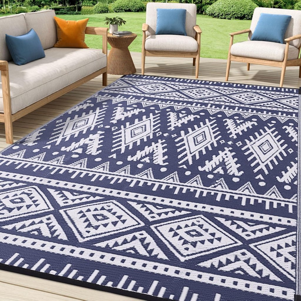 A patio area featuring a light-colored sofa with colorful cushions, two wooden chairs, and a geometric patterned rug in navy blue and white.