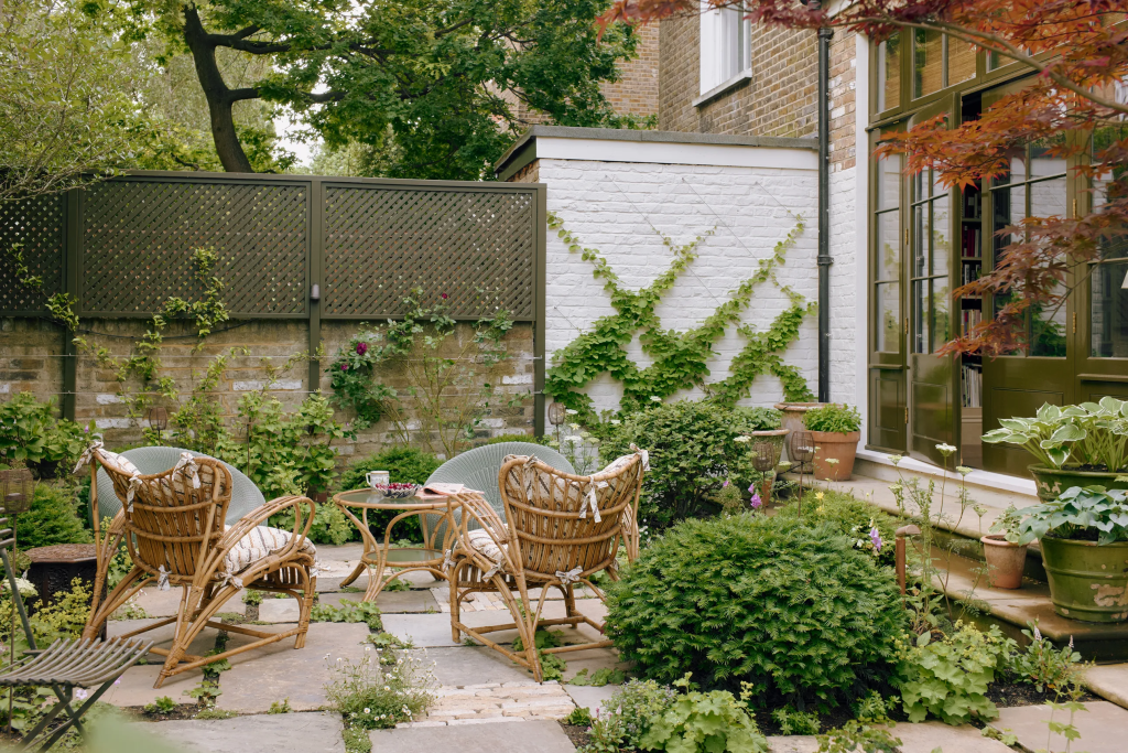 A cozy outdoor seating area featuring two wicker chairs and a small table, surrounded by lush greenery and plants, with a textured wall and window in the background.