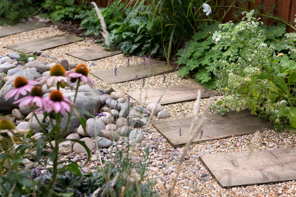 A peaceful garden pathway lined with wooden stepping stones, surrounded by colorful flowers, pebbles, and lush greenery.