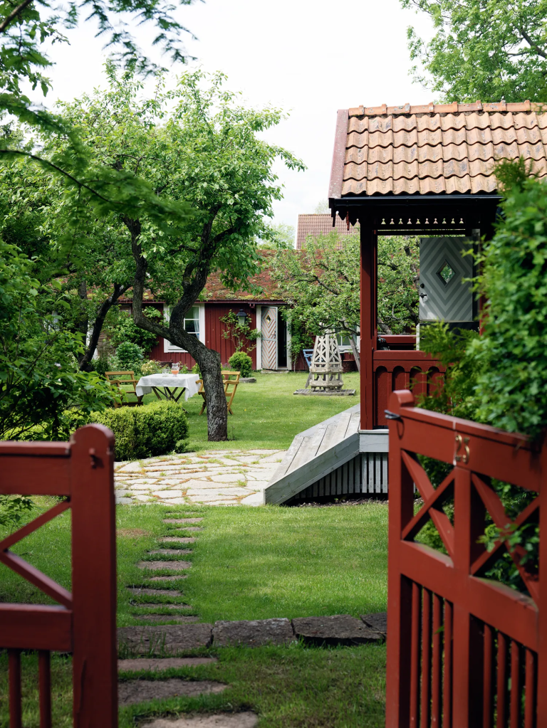 A picturesque garden scene featuring a pathway leading through lush greenery, with a red wooden gazebo and a charming red house in the background.