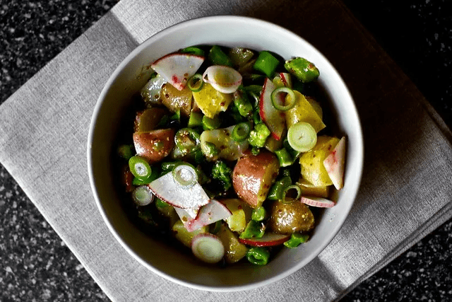 A bowl of spring salad with new potatoes, green peas, sliced radishes, and green onions on a gray napkin, set on a dark surface.