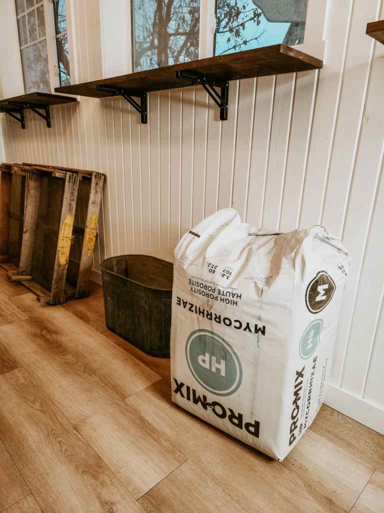 A bag of Promix HP potting soil placed on a wooden floor beside a metal container and wooden shelves.