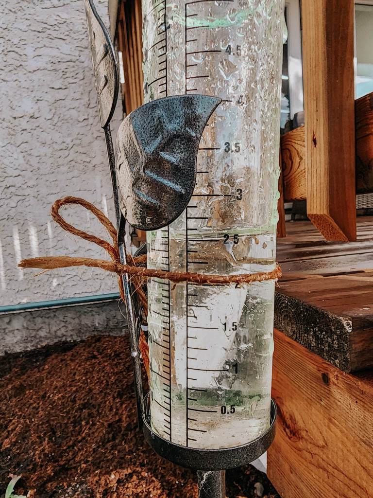 A rain gauge showing accumulated water levels with a decorative holder, placed outdoors near a wooden deck.