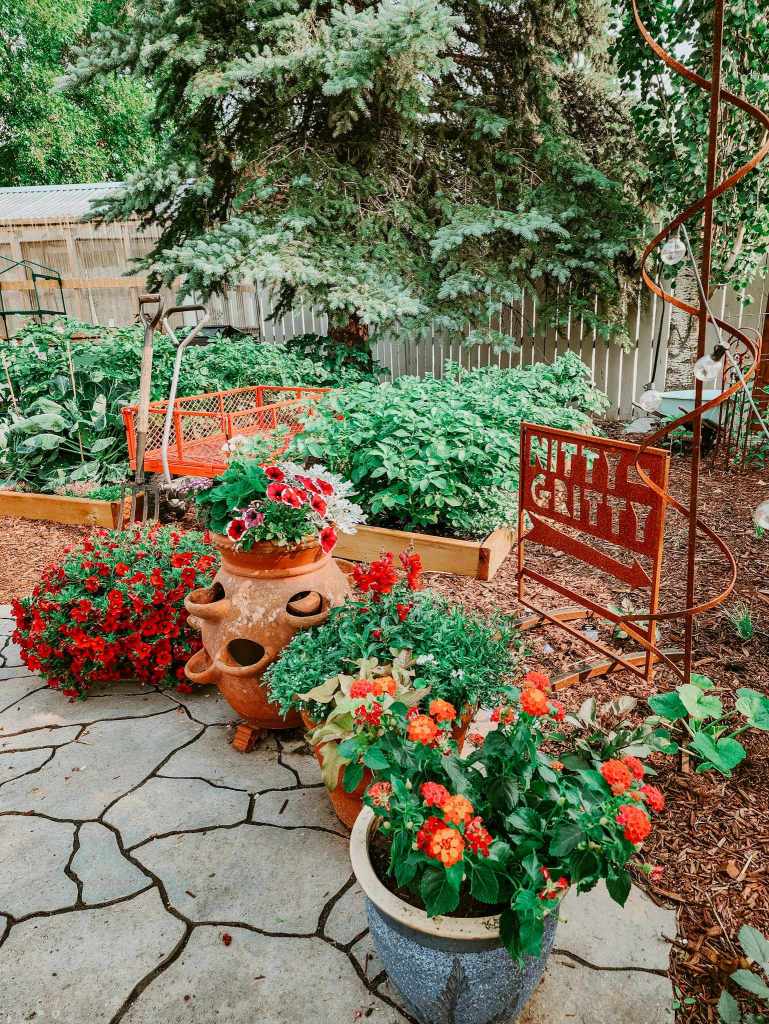 A colorful garden scene featuring various potted plants and flowers, including vibrant red and orange blooms, nestled among green foliage and decorative garden elements. A rustic sign reads 'NITTY GRITTY' in the background.