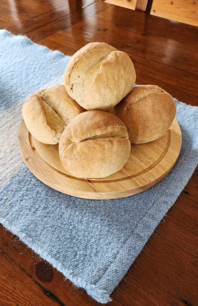 A wooden plate with four freshly baked German bread rolls arranged neatly on a blue woven tablecloth.