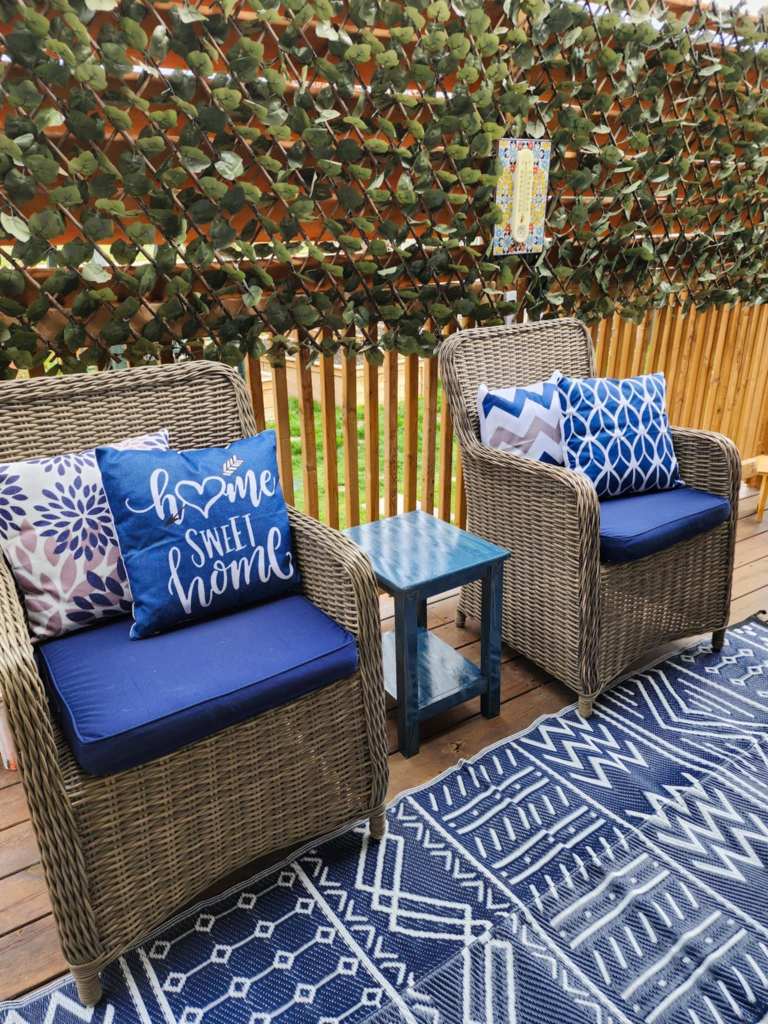 A cozy outdoor seating area featuring two wicker chairs with blue cushions, surrounded by green foliage on a wooden deck, with a decorative rug underneath.
