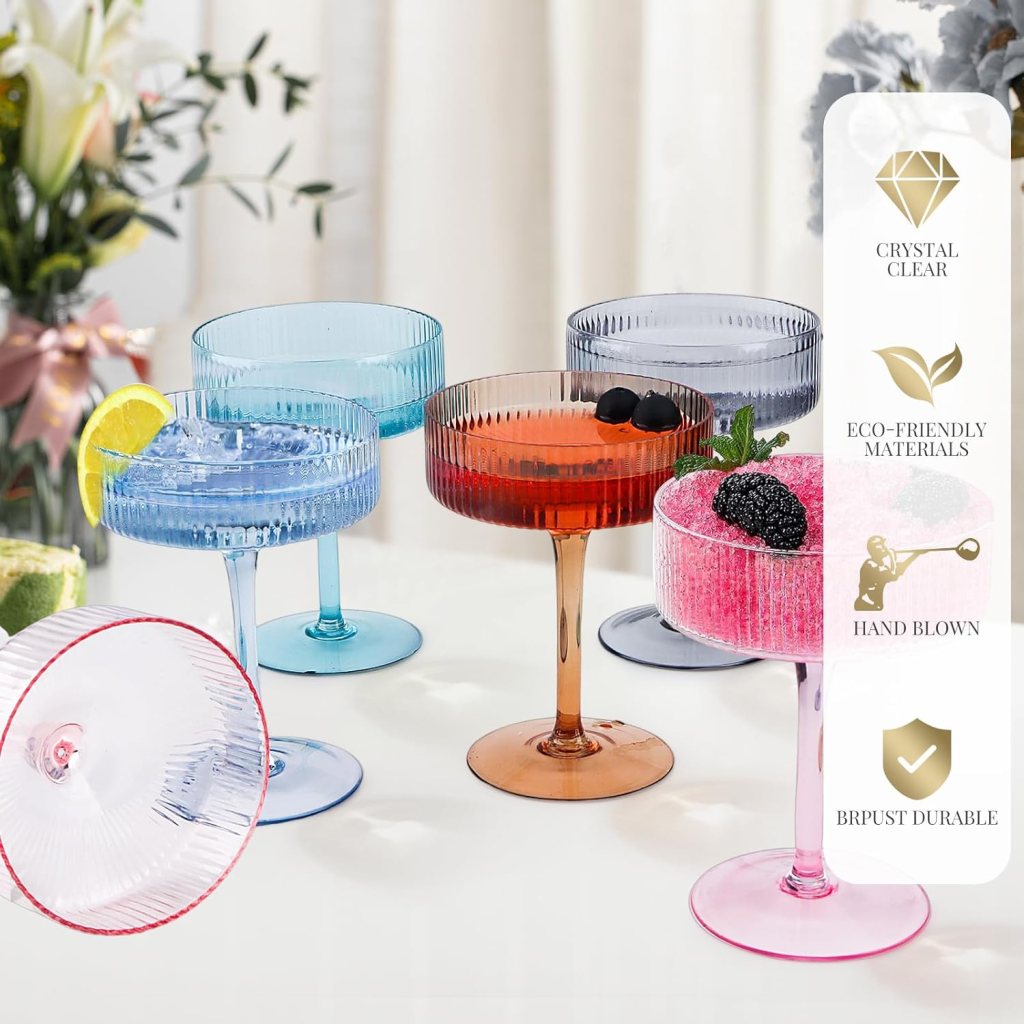 A collection of colorful, hand-blown cocktail glasses in various shades, accompanied by lemon and berry garnishes, displayed on a table with a floral arrangement in the background.