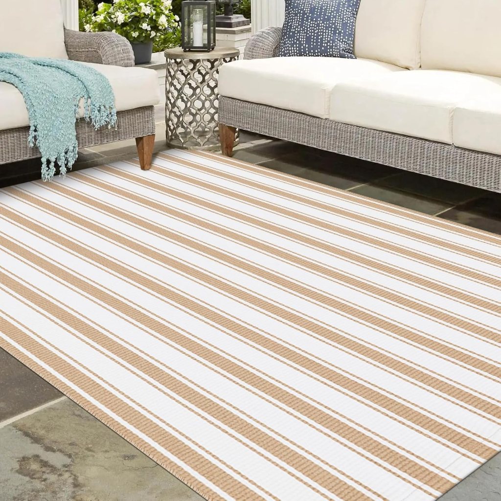 A striped outdoor rug in neutral tones laid on a stone patio, with a comfortable seating area featuring a light-colored sofa and decorative pillows.