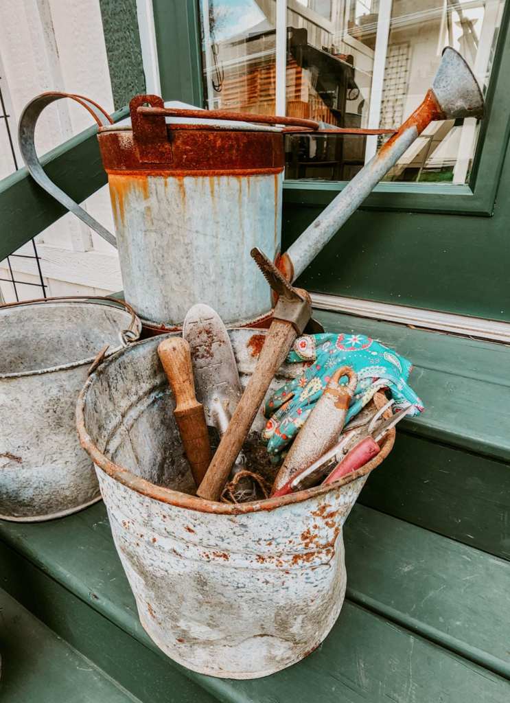 A rustic watering can and gardening tools inside an old metal bucket, placed on a wooden surface.