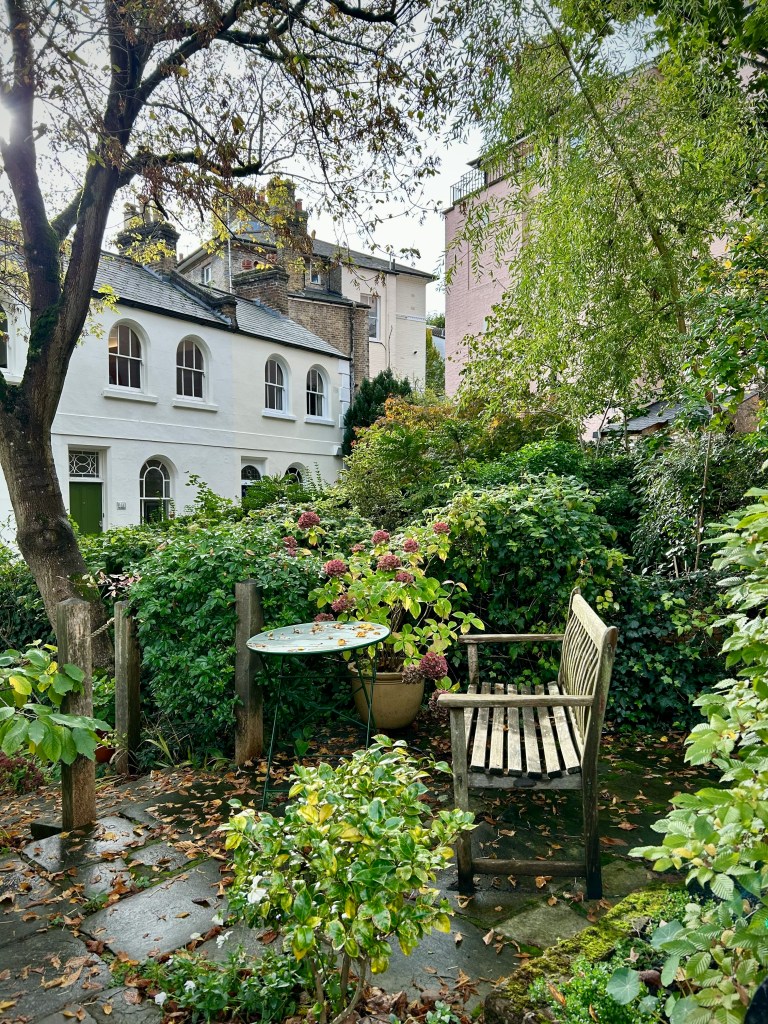 A quaint garden scene featuring a wooden bench and a small round table surrounded by lush greenery and flowering plants, with a backdrop of charming residential buildings.