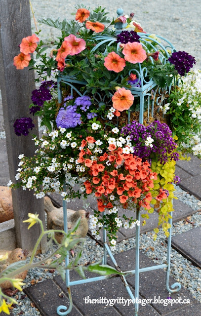 A colorful garden planter overflowing with vibrant flowers, including orange petunias, purple and white calibrachoas, and green foliage, displayed on a decorative metal stand.