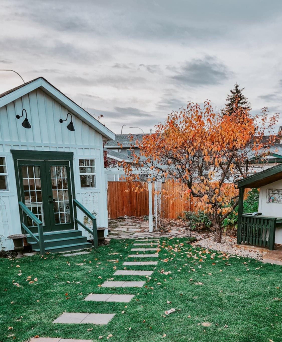 A quaint backyard scene featuring a small white cottage with green accents, a pathway of stone pavers, and a tree with autumn leaves.