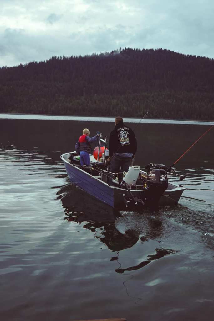 A child and an adult are fishing together from a small boat on a calm lake, surrounded by a dense forested landscape under a cloudy sky.
