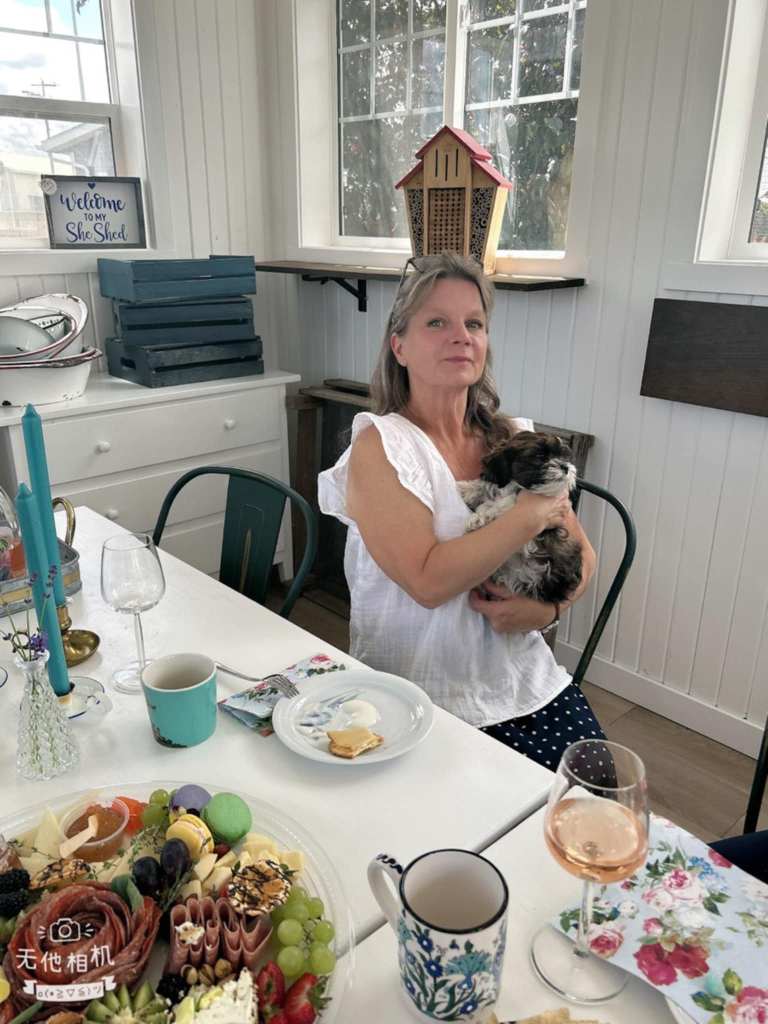 A woman holding a small puppy while sitting at a table with a variety of food, including fruits and drinks, in a bright room with large windows.
