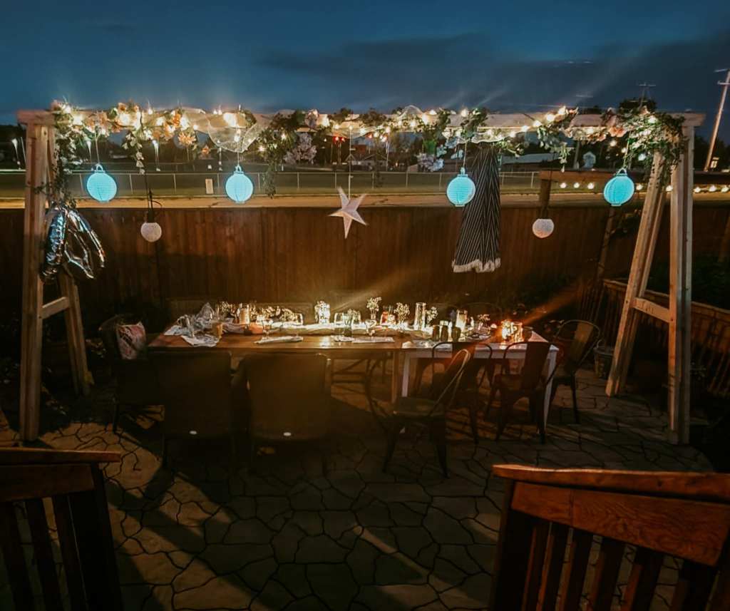 An outdoor dining setup at night with a large table decorated with candles and illuminated by string lights and lanterns, surrounded by wooden chairs.