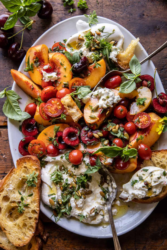 A vibrant platter featuring a colorful mix of peaches, cherries, cherry tomatoes, and fresh herbs, garnished with creamy burrata cheese and toasted bread slices.