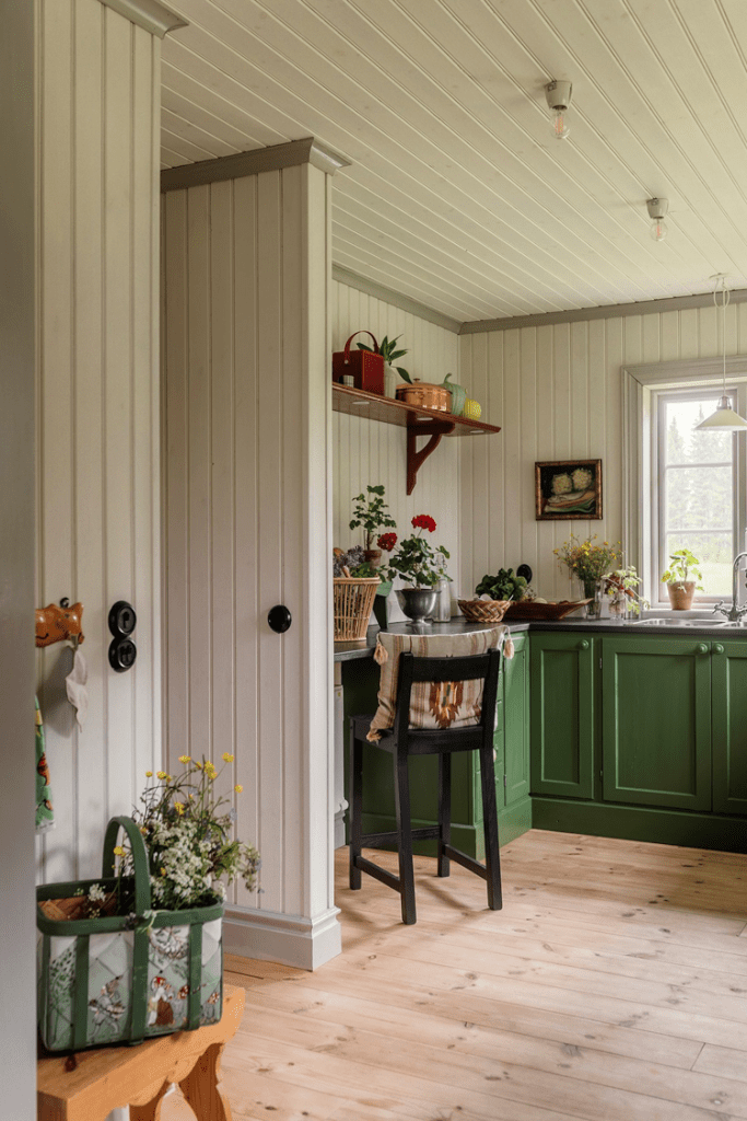 A cozy kitchen featuring green cabinets, wooden flooring, and plants on the windowsill. A small table and chair are positioned near a wall with decorative shelves.