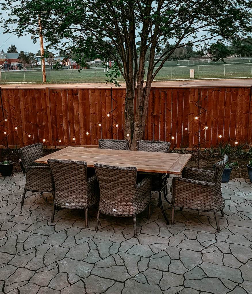 A cozy outdoor dining set with a wooden table and wicker chairs under a tree, situated on a stone patio with decorative string lights in the background.