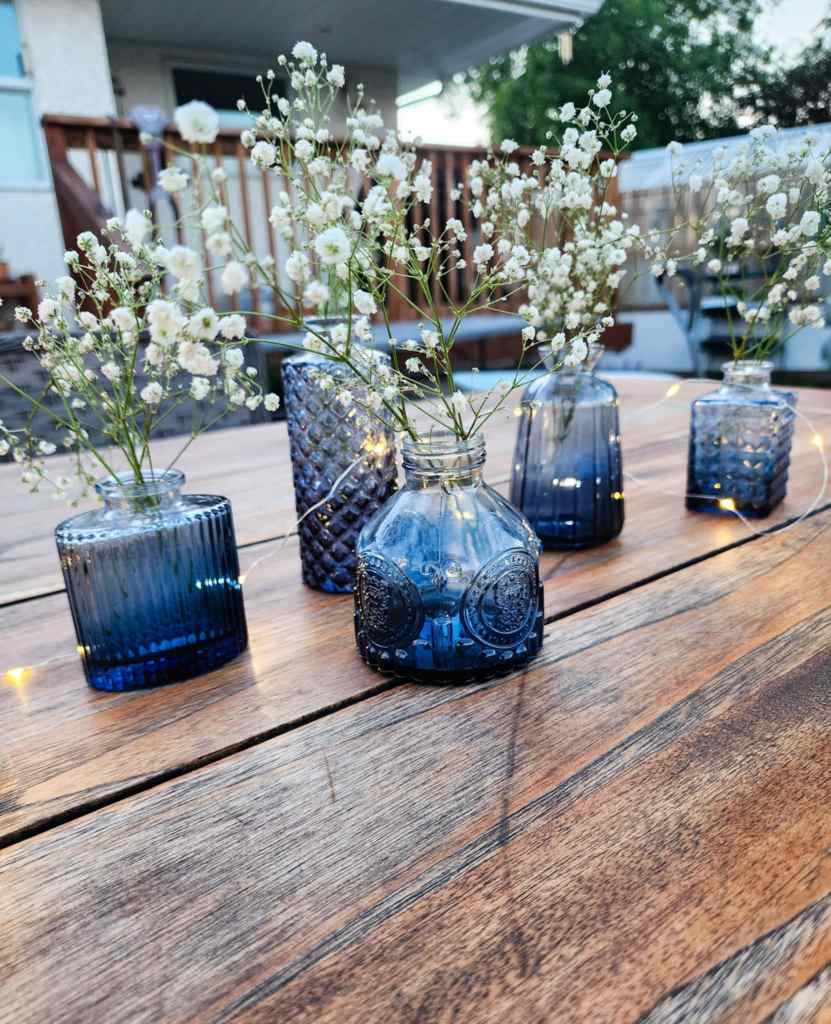 A collection of blue glass vases containing small white flowers arranged on a wooden table, with soft evening light in the background.