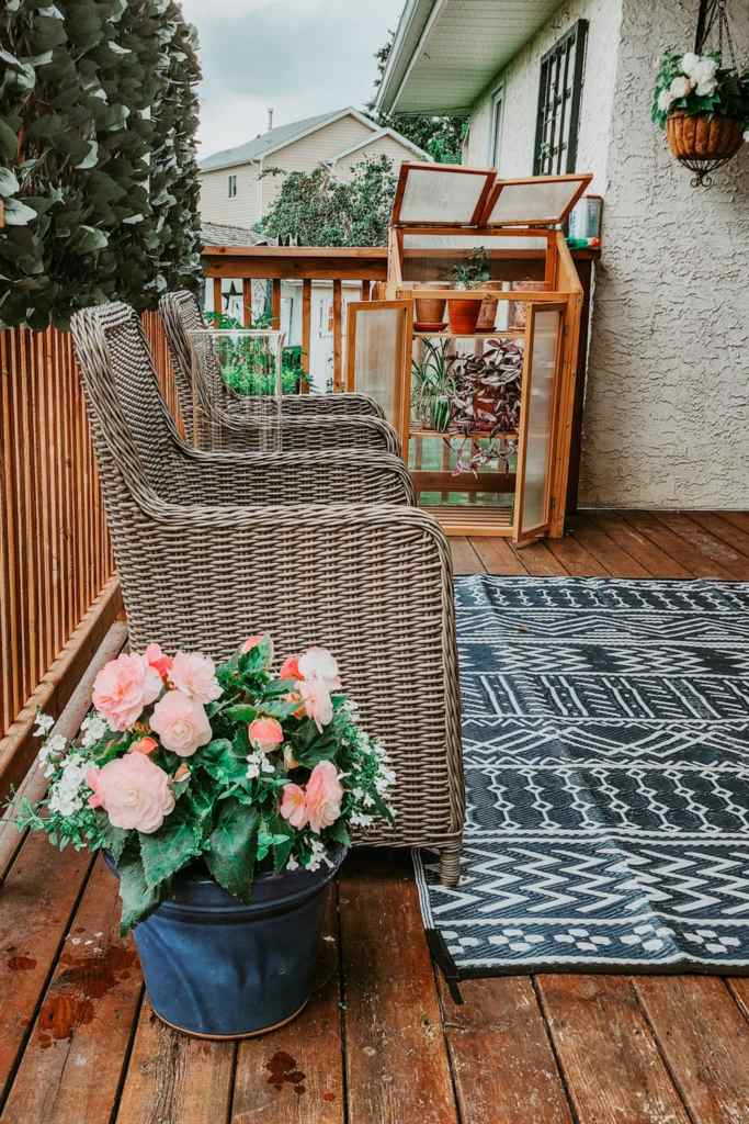 Wicker chairs on a wooden deck with a potted flower arrangement and a small greenhouse.