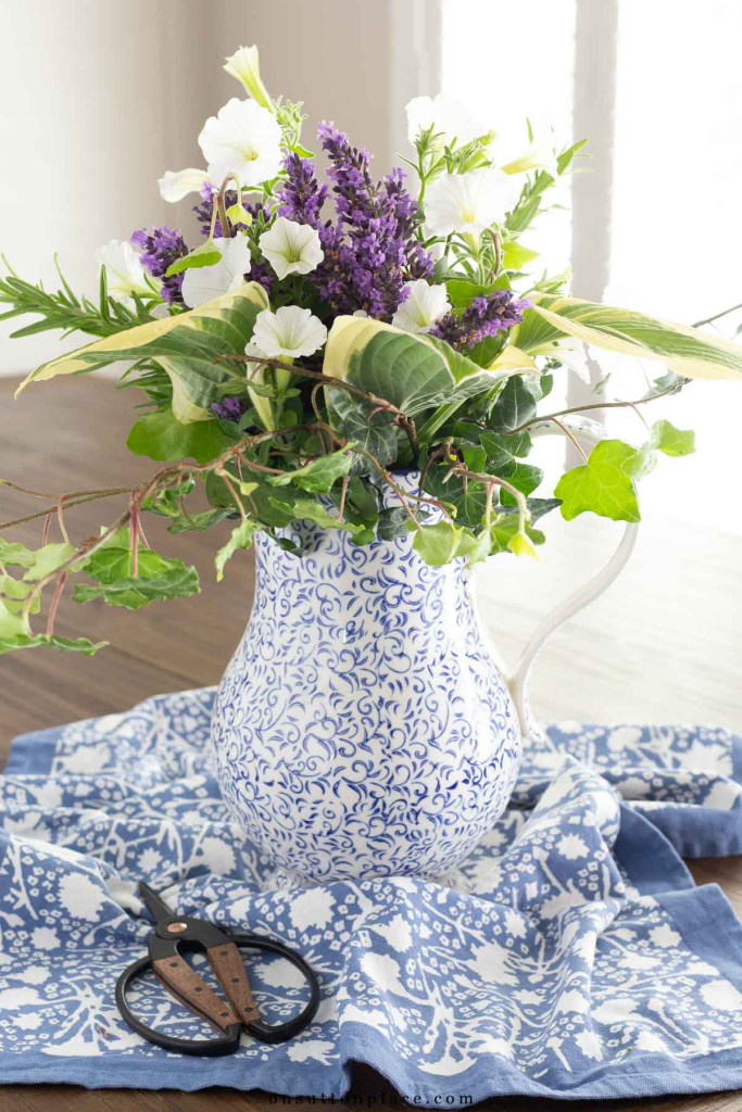 A floral arrangement in a blue and white patterned pitcher, featuring purple flowers and greenery, placed on a blue patterned cloth with pruning scissors nearby.
