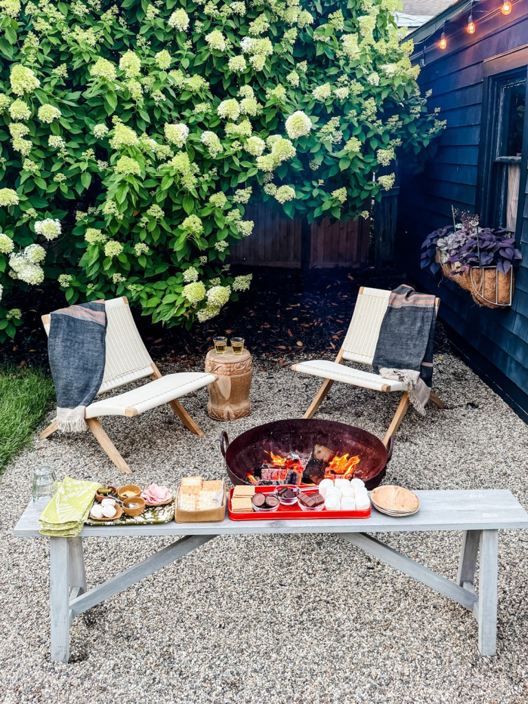A cozy outdoor fire pit setup featuring two chairs with blankets and a wooden table displaying s'mores ingredients, surrounded by lush green hydrangeas.