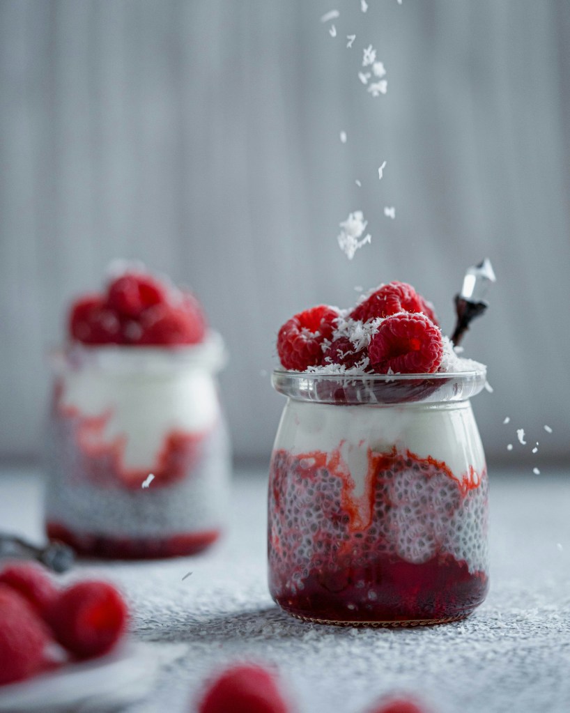 A close-up of jars filled with layers of raspberry puree, yogurt, and chia seeds, topped with fresh raspberries and sprinkled coconut flakes.