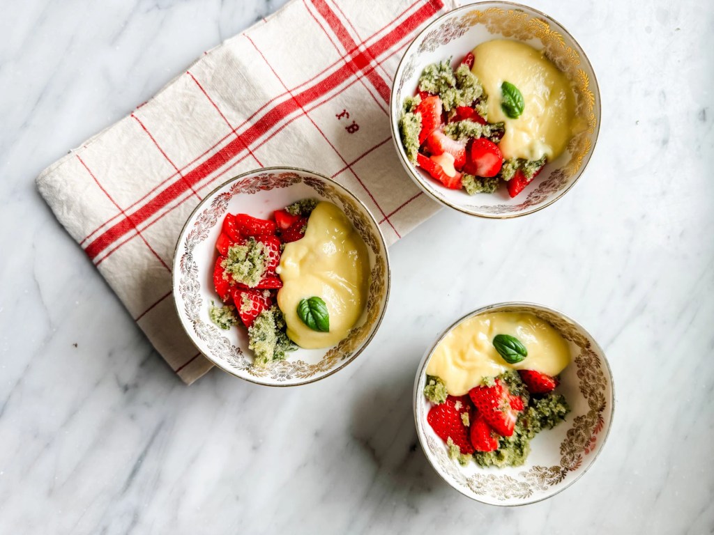 Three decorative bowls filled with strawberries, a creamy yellow dessert, and crumbled green topping, placed on a marble surface with a red and white checkered cloth.