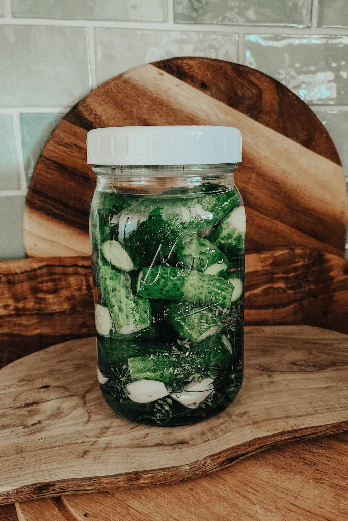 A jar filled with cucumber slices, garlic cloves, and fresh herbs submerged in pickling brine, resting on a wooden surface.