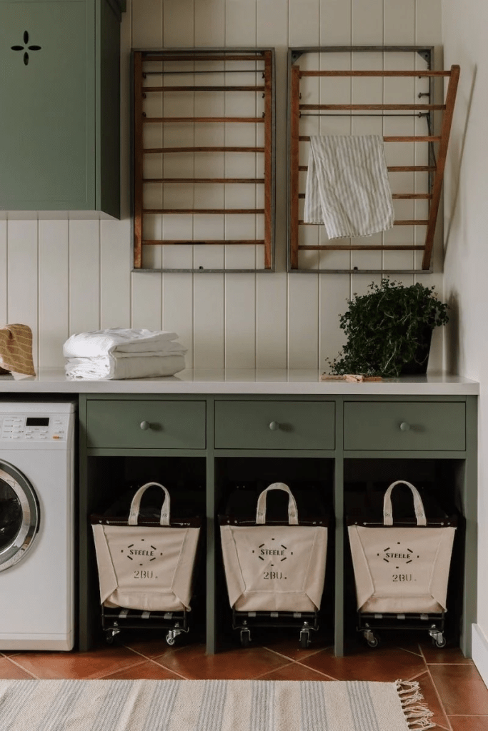 A functional laundry room featuring green cabinets, a washing machine, and rolling laundry baskets underneath a countertop, with wooden drying racks mounted on the wall.