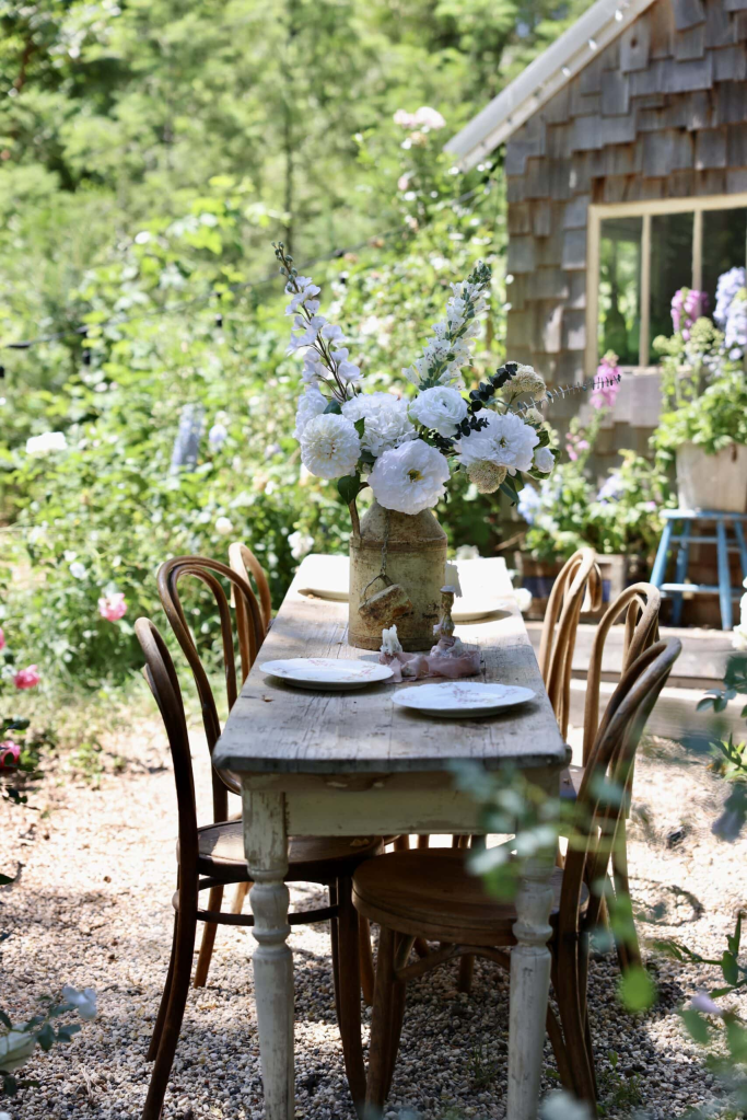 A rustic outdoor dining table set with plates, surrounded by lush green plants and flowers, with a charming wooden shed in the background.