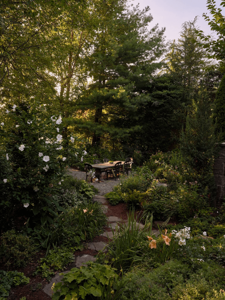 A serene garden scene featuring a rustic dining table surrounded by lush greenery, colorful flowers, and tall trees under soft evening light.