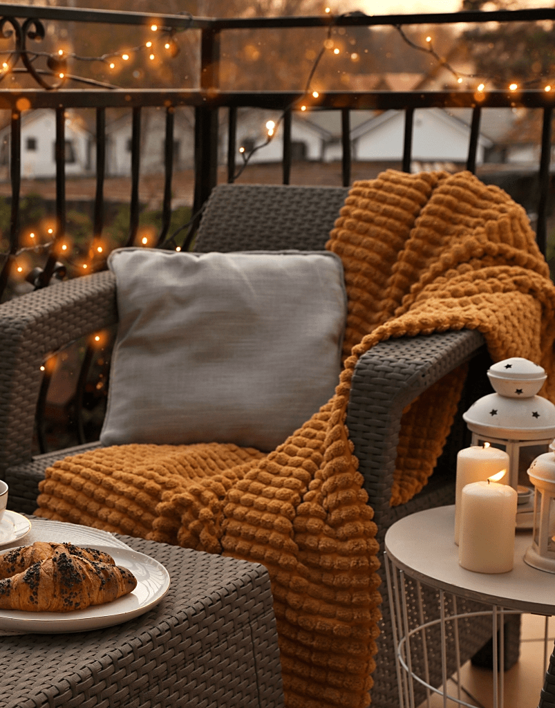 A cozy outdoor seating area featuring a grey woven armchair draped with a mustard-colored blanket, a light grey cushion, and a table with a plate of croissants and a warm drink, surrounded by soft glowing fairy lights.