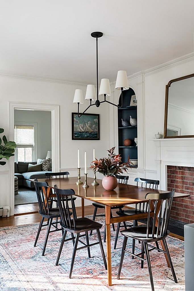 A cozy dining room featuring a wooden table surrounded by black chairs, with a minimalist chandelier above, a colorful rug underneath, and a plant nearby.