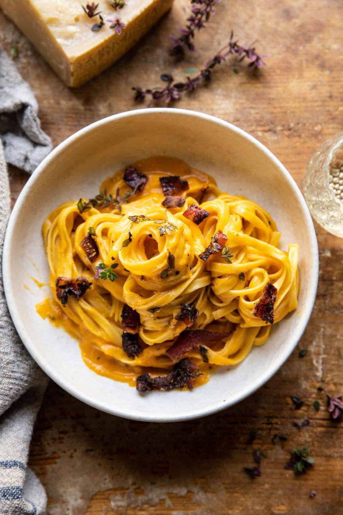 A bowl of creamy pasta with roasted butternut squash sauce, garnished with crispy bacon pieces and fresh thyme, served alongside a wedge of cheese.