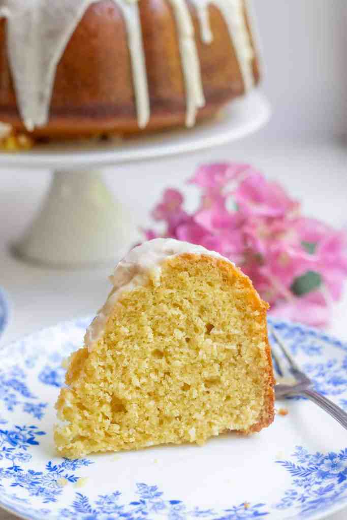 A slice of cake with a glossy icing on a blue floral plate, accompanied by a fork and some pink flowers in the background.