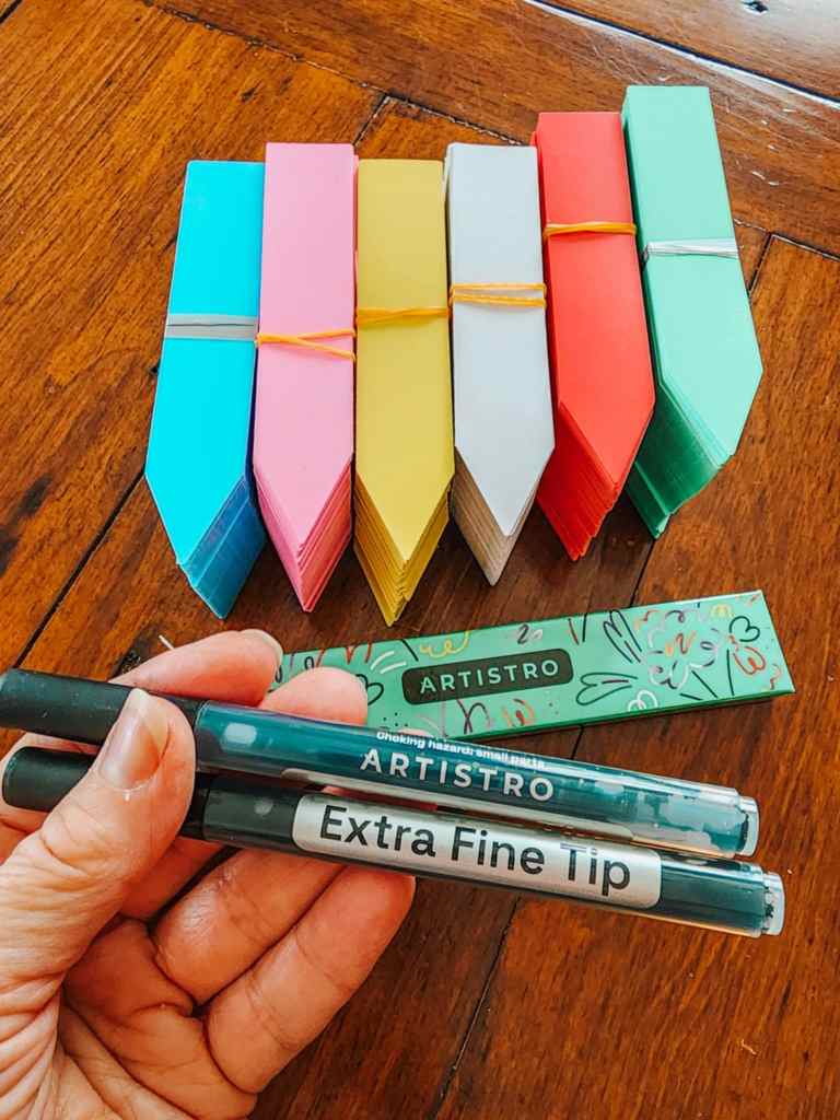 A hand holding two marking pens and a pack of colorful plant labels arranged vertically.