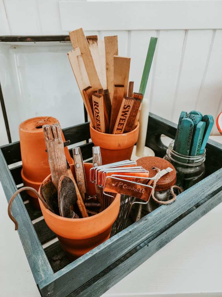 A collection of gardening tools, including labeled wooden plant markers, stored in terracotta pots and a wooden crate, showcasing organization for gardening tasks.