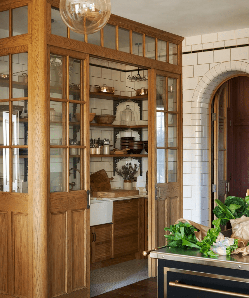 An open kitchen area featuring wooden double doors, glass panels, and shelving with various kitchen items, including bowls and utensils. Fresh greens and groceries are visible on a countertop.