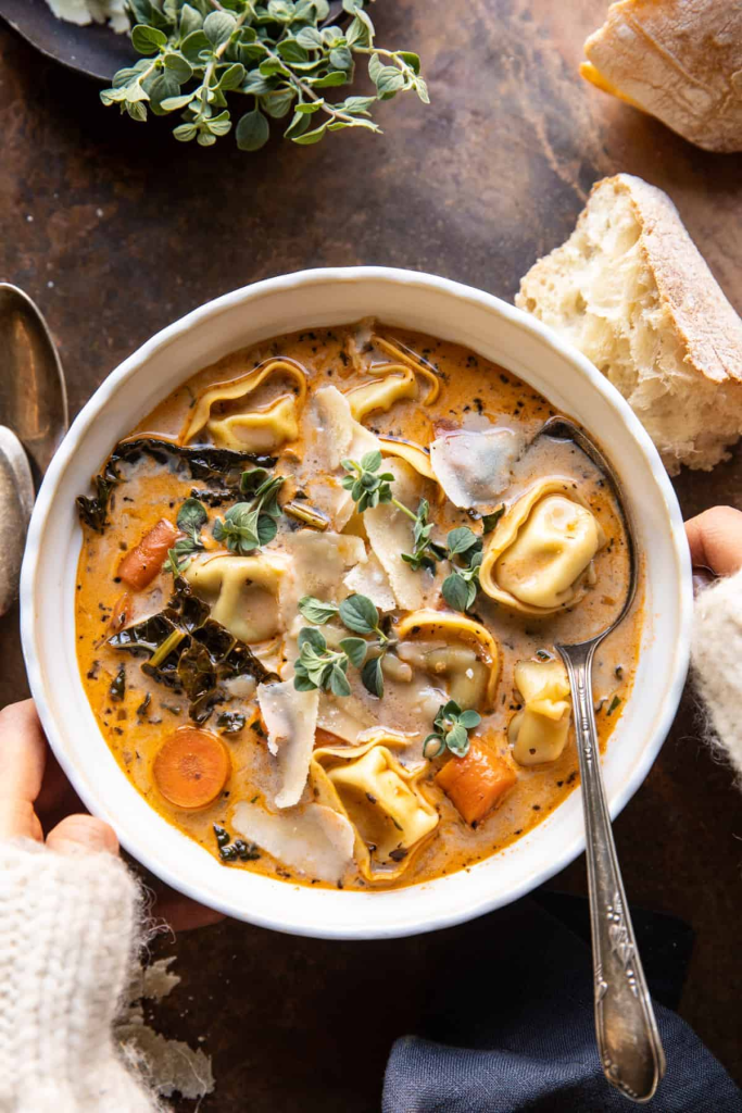 A warm bowl of creamy tortellini soup garnished with fresh herbs, accompanied by pieces of bread, resting on a rustic table.