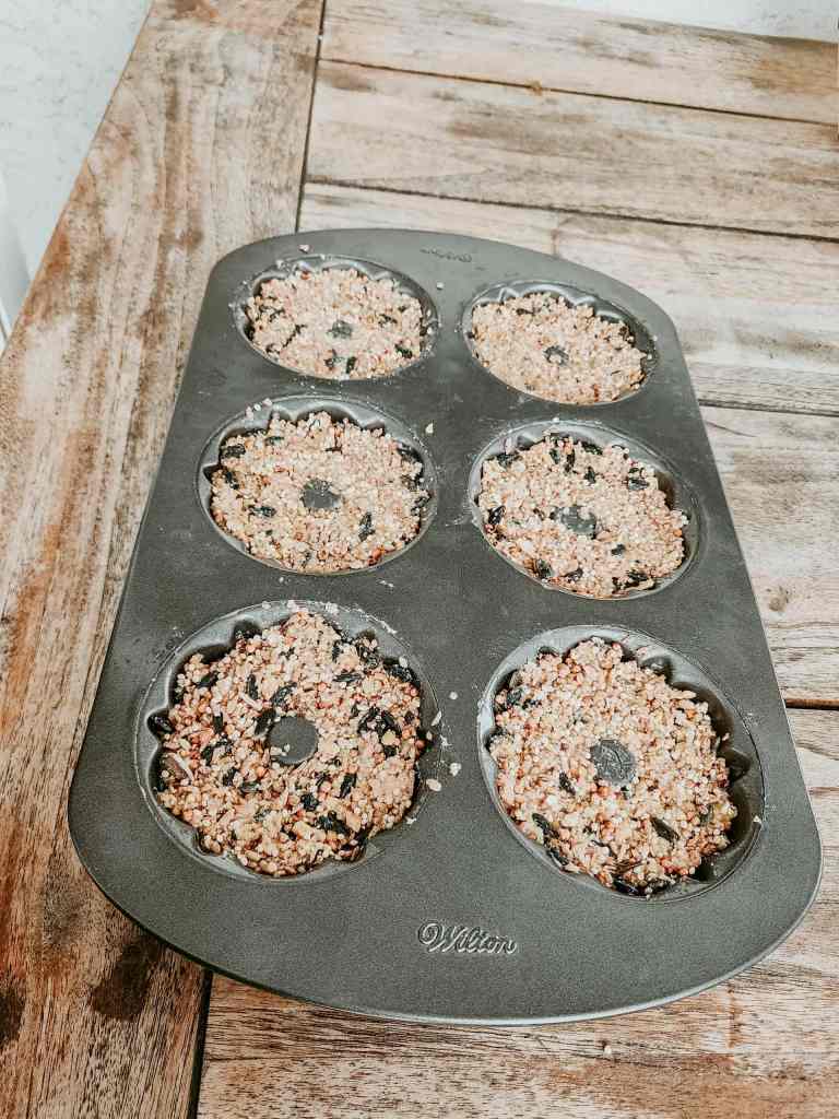 A mini bundt cake pan filled with birdseed mixture, set on a wooden surface, showing six birdseed wreaths in the mold.