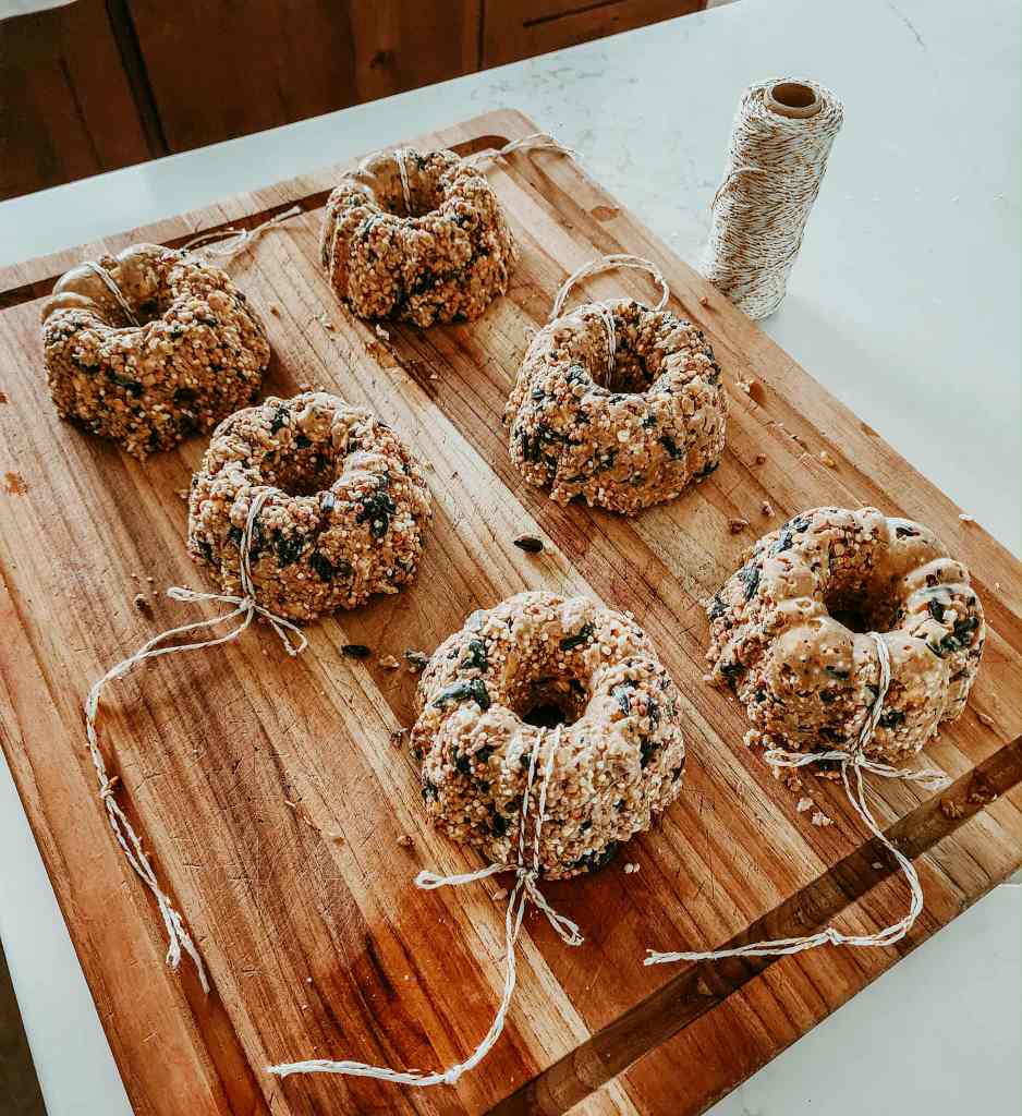 A wooden cutting board displaying six mini birdseed wreaths with twine for hanging, alongside a roll of twine.