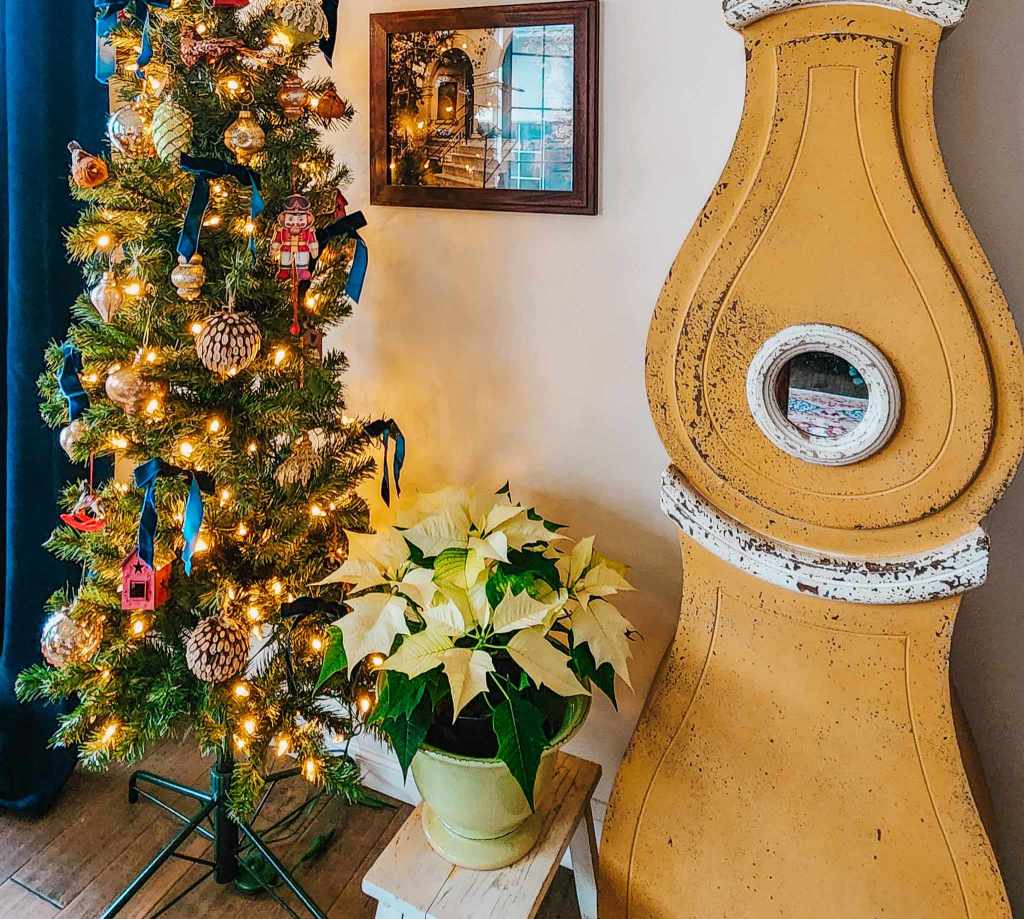 A decorated Christmas tree with lights and ornaments next to a pot of white poinsettias, beside a vintage-style yellow decorative piece on a small table.