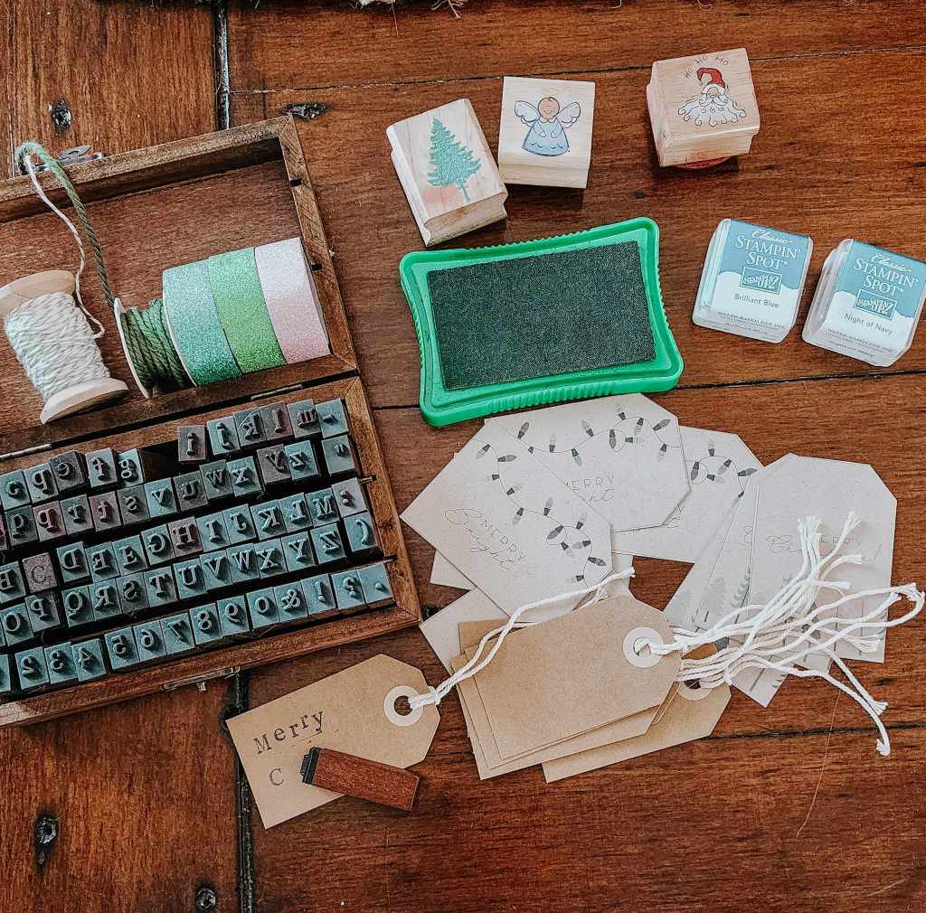 A flat lay of crafting supplies including a wooden box of alphabet stamps, spools of ribbon, ink pads, and decorative gift tags on a wooden surface.