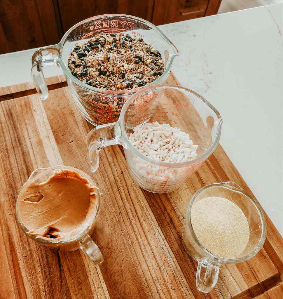 Ingredients for birdseed wreaths arranged on a wooden cutting board, including birdseed mix, peanut butter, cornmeal, and additional grain.