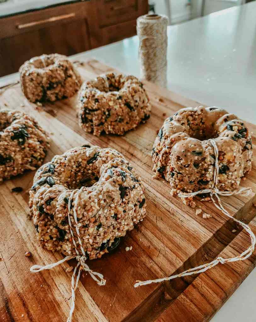 Several mini birdseed wreaths made from seeds and nuts, tied with twine, displayed on a wooden cutting board.